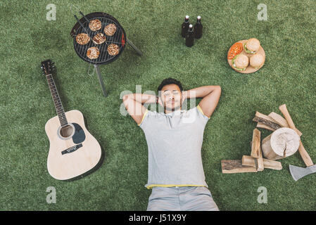 Vue du dessus de l'heureux jeune homme se reposant sur l'herbe avec guitare et de l'alimentation pour des pique Banque D'Images