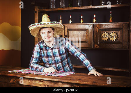 Smiling man comme barman dans un sombrero se pencha sur comptoir de pub mexicain, une étagère avec des sauces épicées et des bouteilles vides dans l'arrière-plan Banque D'Images