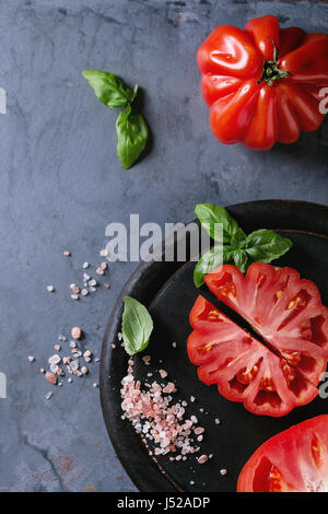 Ensemble et les tranches de tomates Coeur de boeuf bio. Tomate coeur de boeuf avec du sel rose et basilic sur planche en bois noir sur gris bleu metal textur Banque D'Images