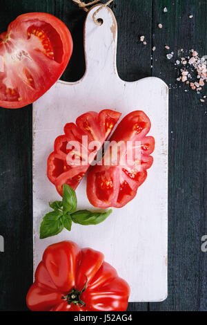 Ensemble et les tranches de tomates Coeur de boeuf bio. Tomate coeur de boeuf avec du sel rose et basilic sur planche en bois blanc sur fond de bois sombre. Banque D'Images