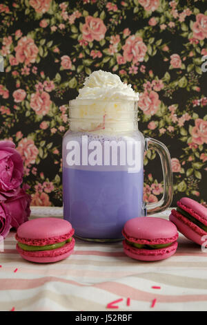 Café pourpre en pot Mason Tasse stylisée avec ses macarons et les roses et une décoration colorée sur fond de motif floral. Lait frappé aux bleuets. Licorne Banque D'Images