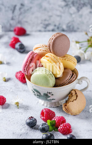 Variété de desserts sucrés macarons français colorés avec différents fourrages servi en tasse de thé vintage avec fleurs de printemps et de baies fraîches sur te gris Banque D'Images