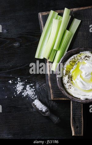 Céleri frais avec du yaourt et l'huile d'olive dans Bol en céramique, servi avec du sel de mer sur bois planche à découper en bois noir sur fond brûlé. Vue supérieure w Banque D'Images