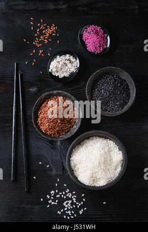 Variété assortiment de matières colorées non cuit le riz blanc, noir, marron, rose en noir sur fond de bois brûlé bols avec des baguettes. Vue supérieure avec Banque D'Images