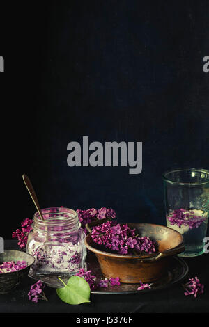 Pot en verre de lilas fleurs en sucre, verre de l'eau avec du citron et de lilas lilas en fleurs fraîches vintage bol en cuivre sur nappe noir sur noir. Da Banque D'Images