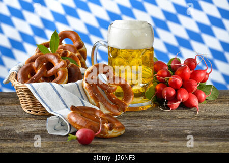 Bretzels frais dans un grenier, un bouquet de radis et une bière bavaroise chope sur une table en bois, dans l'arrière-plan le drapeau blanc-bleu de Bavière Banque D'Images