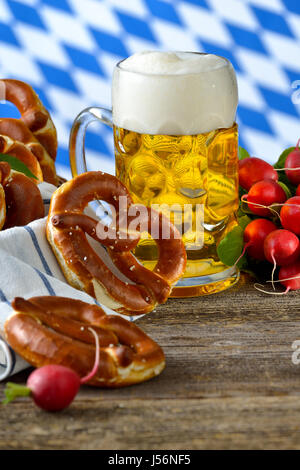 Bretzels frais dans un grenier, un bouquet de radis et un litre de bière bavaroise sur une table en bois, dans l'arrière-plan le drapeau blanc-bleu de Bavière Banque D'Images