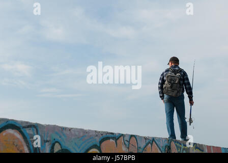 Homme adulte avec une canne à pêche et un sac à dos va à la pêche à pied sur le mur de graffiti Banque D'Images