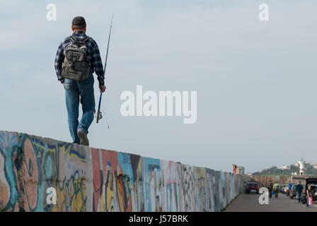 Homme adulte avec une canne à pêche et un sac à dos va à la pêche à pied sur le mur de graffiti sur fond de gens et de voitures et le phare Banque D'Images