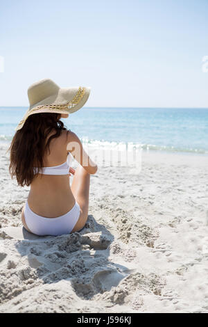 De vie arrière woman wearing sun hat while sitting on sand at beach Banque D'Images