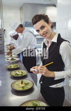 Portrait of smiling waitress avec bloc-notes de cuisine commerciale et chefs preparing food in background Banque D'Images