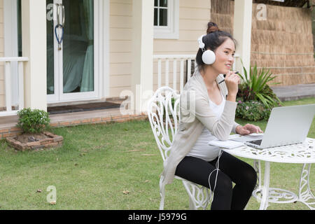 Young Asian woman sitting on chair d'écouter de la musique et à l'aide d'un ordinateur portable. Belle femme attendent à avoir l'enfant reste dans un jardin en face de il Banque D'Images