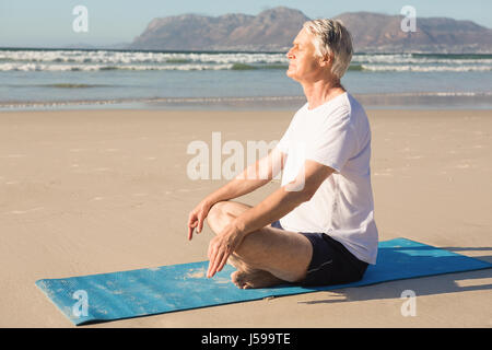 Vue latérale du senior man meditating on mat at beach Banque D'Images