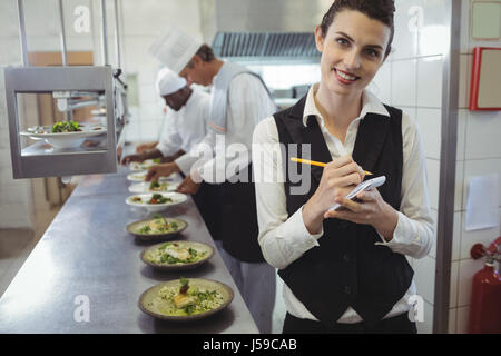 Portrait of smiling waitress avec bloc-notes de cuisine commerciale et chefs preparing food in background Banque D'Images