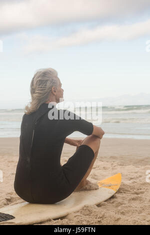 Vue arrière du senior woman sitting on surfboard contre ciel nuageux Banque D'Images