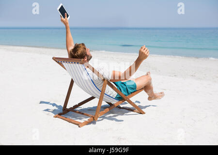 Man with arms outstretched holding digital tablet, assis sur une chaise longue à beach Banque D'Images