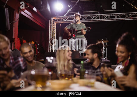 Smiling friends sitting by table avec le musicien en arrière-plan at nightclub Banque D'Images
