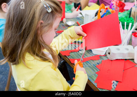 Petite fille à l'aide de ciseaux pour couper les formes d'un morceau de papier rouge. Elle est dans une classe de maternelle avec les autres étudiants. Banque D'Images