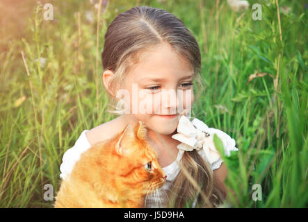 Cute little girl holding est un chat roux dans la nature assis dans l'herbe. Banque D'Images
