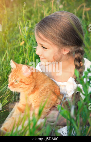 Cute little girl holding est un chat roux dans la nature assis dans l'herbe. Banque D'Images