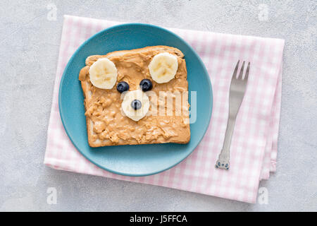 Ours toast pour les enfants. Beurre d'arachide croquant, banane et myrtille toast avec mignon ours visage sur une plaque bleue sur textile à carreaux rose. À Banque D'Images