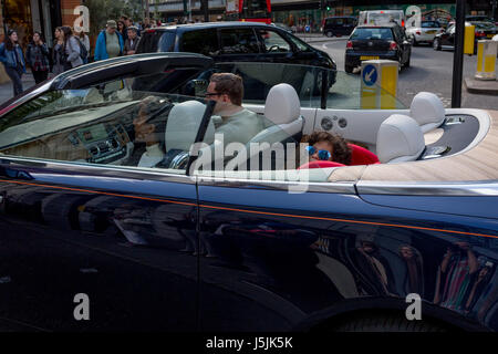 De riches adultes et un enfant portant des lunettes de soleil qui sourit à l'arrière d'une voiture à toit ouvert qui reste immobile aux feux de circulation dans la région de Sloane Square, le 14 mai 2017, à Londres, en Angleterre. Banque D'Images