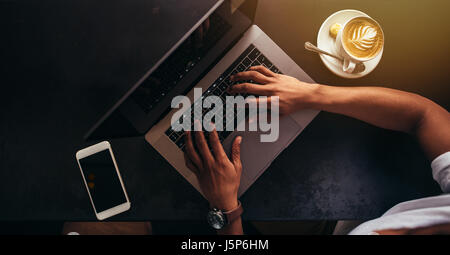 Close up shot of young man sitting at coffee shop et le travail sur ordinateur portable. Se concentrer sur les mains de la saisie sur clavier d'ordinateur portable avec téléphone mobile et tasse de café Banque D'Images