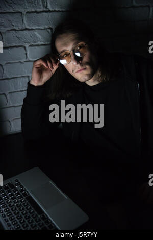 Image de jeune homme concentré portant des lunettes à l'aide d'un ordinateur portable à la maison à l'intérieur de nuit. En regardant la caméra. Banque D'Images