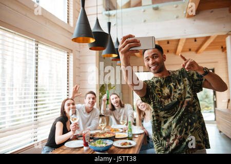 Jeune homme africain joyeux selfies en tenant avec ses amis à la table à la maison Banque D'Images