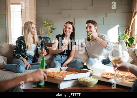 Groupe de jeunes amis joyeux avec la pizza, vin et bière à parler et s'amuser sur canapé à la maison Banque D'Images