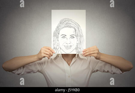 Belle femme avec happy self portrait en face de son visage, cachant ses vraies émotions, l'humeur, isolé sur fond de mur gris. La vie privée, l'identit Banque D'Images