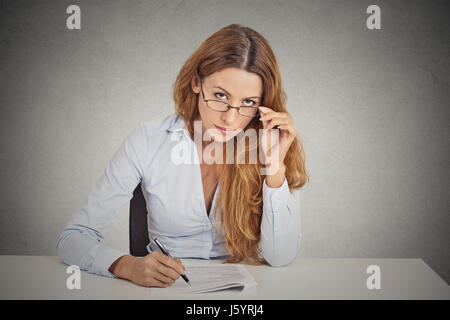 Les curieux businesswoman with glasses sitting at desk avec scepticisme en vous regardant l'examen isolé sur fond de Mur Gris bureau. Les droits de fa Banque D'Images