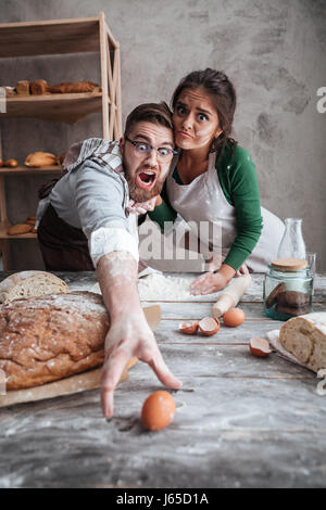 Jeune homme et femme choqué essaye d'attraper l'œuf sur table avec la farine en cuisine gris Banque D'Images