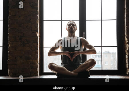 Portrait d'un jeune sportif calme méditant dans lotus posent, assis sur le rebord de la fenêtre dans la salle de sport Banque D'Images