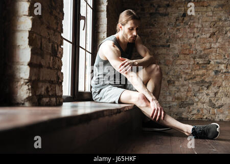 Portrait d'un jeune homme souffrant d'une remise en forme à la jambe alors qu'il était assis sur le rebord de la fenêtre dans la salle de sport Banque D'Images