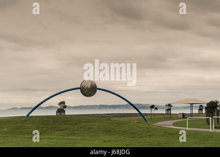 Napier, Nouvelle-Zélande - mars 9, 2017 : Metal écliptique la sculpture de David Trubridge à Beach park sous un ciel de tempête brun. L'océan Pacifique et la verte Banque D'Images