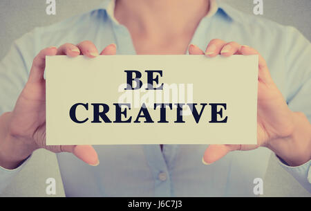 Libre de businesswoman hands holding white card signe avec être créatifs message texte isolé sur fond de bureau mur gris. Image retro style instagram Banque D'Images