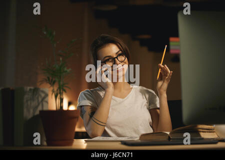 Photo de young smiling woman sitting designer intérieur à conversation de nuit par téléphone mobile et la tenue d'un crayon. À côté. Banque D'Images