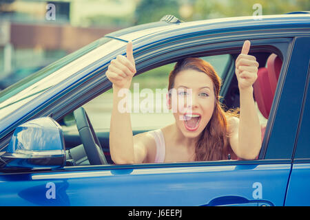 Vue sur la porte latérale conducteur happy smiling woman showing Thumbs up assis à l'intérieur bleu nouvelle voiture à l'extérieur sur l'arrière-plan de stationnement. Belle jeune femme happ Banque D'Images