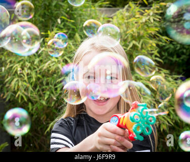 Dix ans fille blonde utilise une petite machine à faire des bulles à l'appareil photo dans un jardin Banque D'Images