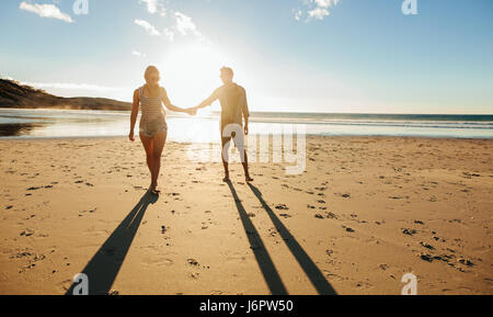 Tourné en plein air romantique de jeune couple en train de marcher le long de la côte se tenant la main. Jeune homme et femme marche sur la plage ensemble au coucher du soleil.