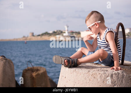 Penser petit Garçon à lunettes est assis sur un brise-lames en béton contre le fond de la mer et le rivage avec lighthouse Banque D'Images