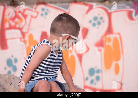 Petit garçon portant des lunettes de soleil assis sur un mur en pierre avant de graffitis tournant autour de Banque D'Images