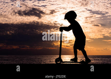 Silhouette d'un little boy wearing helmet riding scooter dans le contexte d'un coucher de soleil sur la mer Banque D'Images