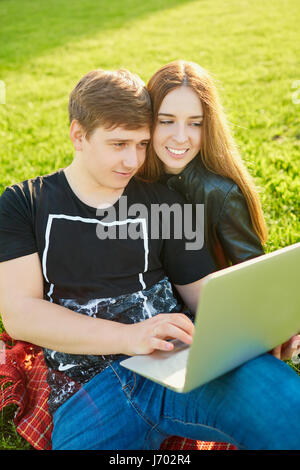 Un couple aimant avec un ordinateur portable dans le parc au coucher du soleil. Banque D'Images