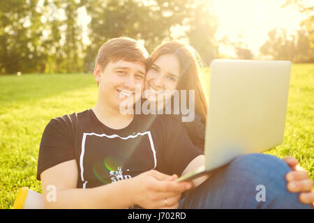 Un couple aimant avec un ordinateur portable dans le parc au coucher du soleil. Banque D'Images