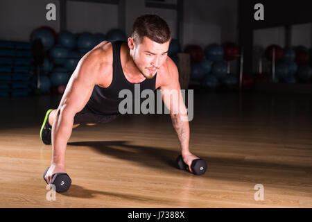 Jeune homme sportif faisant pompes avec haltères courtes dans le cadre de la formation de culturisme. Banque D'Images