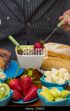 Jeune homme tenant avec une fourchette tremper une fraise recouverte d'une fondue au chocolat à l'intérieur d'un bol blanc avec fruits assortis dans des bols sur la table en bois Banque D'Images