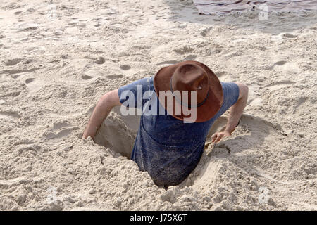 Un jeune homme tente de creuser lui-même hors d'un trou dans le sable qu'il les a en lui-même sur une plage un jour ensoleillé Banque D'Images