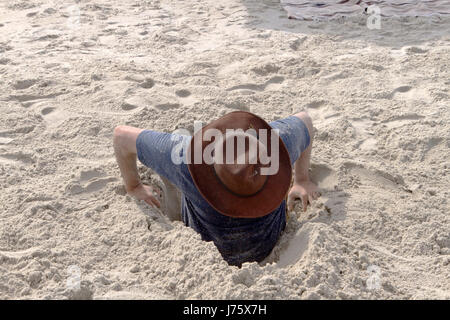 Un jeune homme tente de creuser lui-même hors d'un trou dans le sable qu'il les a en lui-même sur une plage un jour ensoleillé Banque D'Images